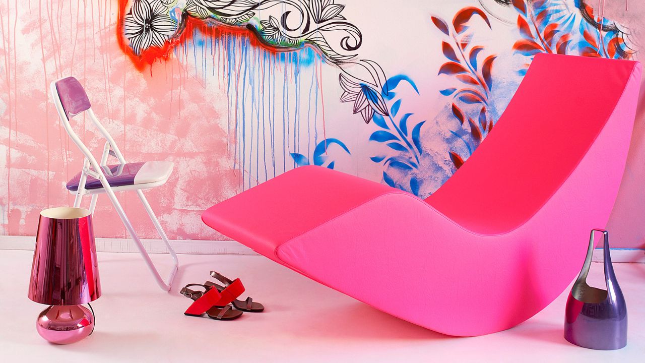 Vibrant, graffiti-inspired pink wall with a pink lounge chair, a small white chair, a purple and pink lamp, and sandals.