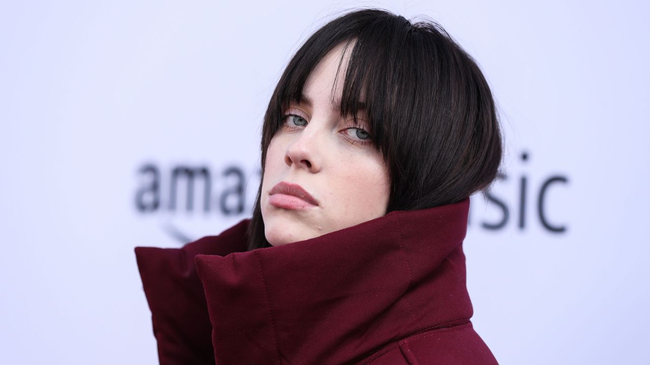 Billie Eilish dark hair and a serious expression, wearing a maroon coat, outdoors.