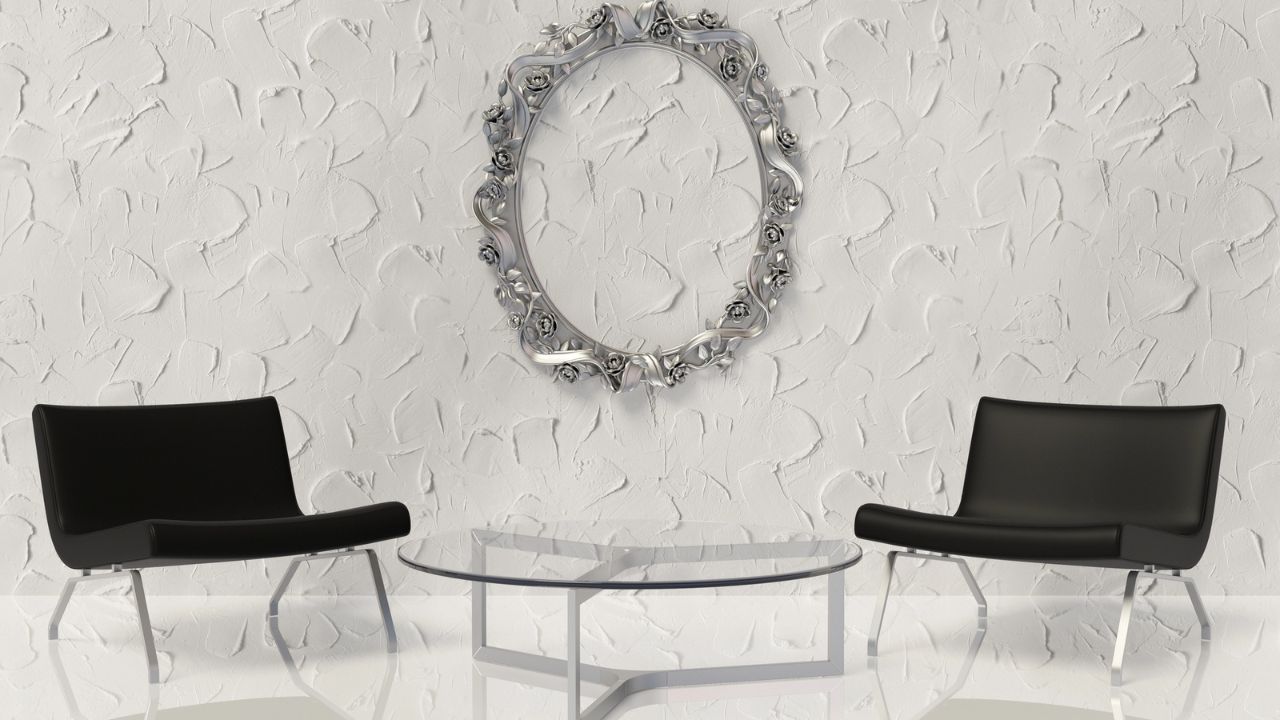 Two black chairs with a glass table in the middle, against a textured white wall with a decorative mirror.