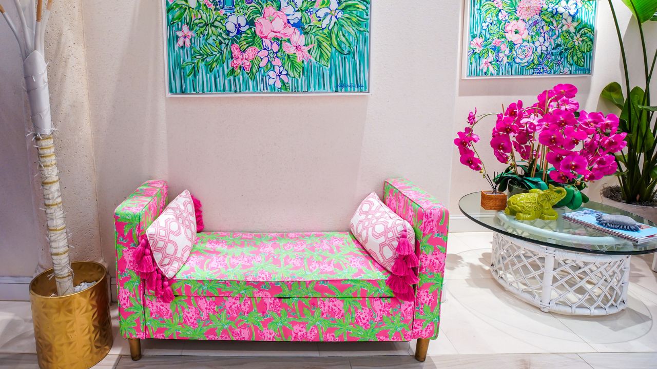 Small pink floral paintings above a colorful pink and green upholstered bench with tassel pillows and vibrant plants.