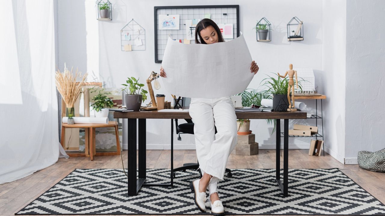 A woman sits on a desk reading large papers in a stylish, plant-filled office.