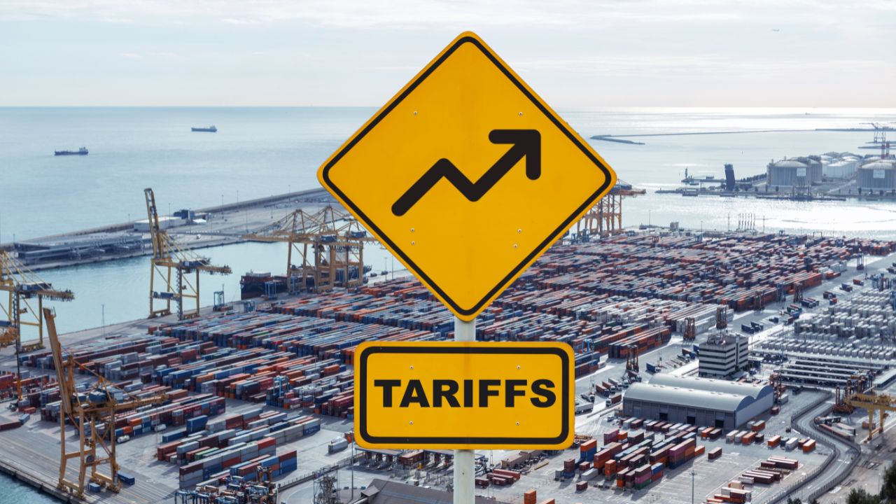 A yellow road sign with an upward arrow and the word "TARIFFS" at the bottom, overlooking a port with containers.