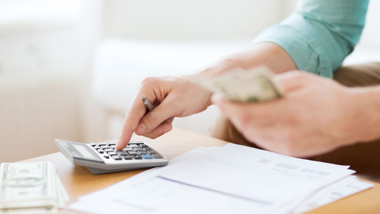 A person using a calculator and making notes on a desk with papers and money.