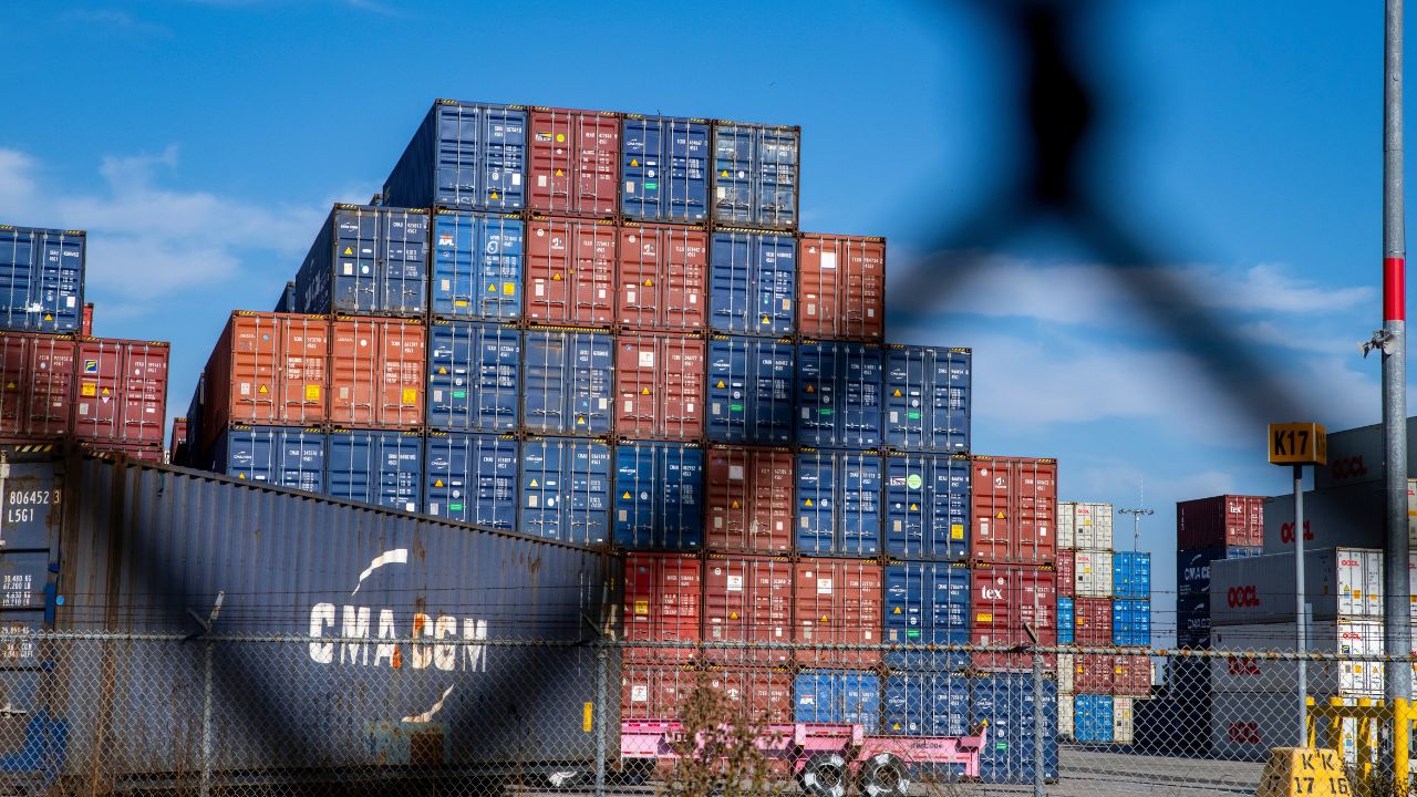 A delivery of stacked shipping containers at a port with cranes in the background.