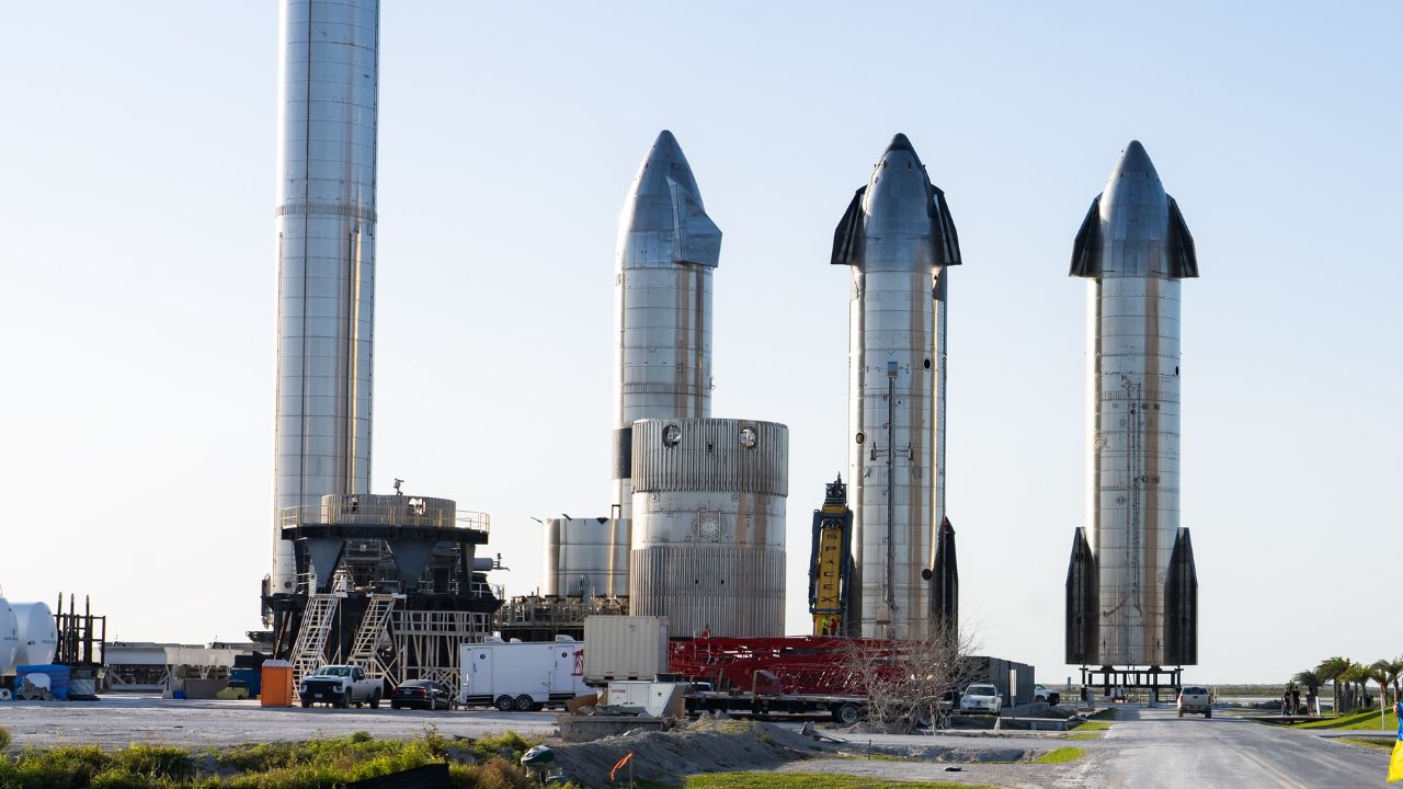 A row of SpaceX Starship rockets standing at the launch site in Boca Chica, Texas. The rockets are towering, with shiny metal exteriors, ready for testing or launch preparations.