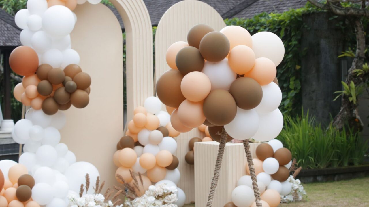 A party stage setup with white. brown, and beige balloon clusters and a warm arched backdrop.
