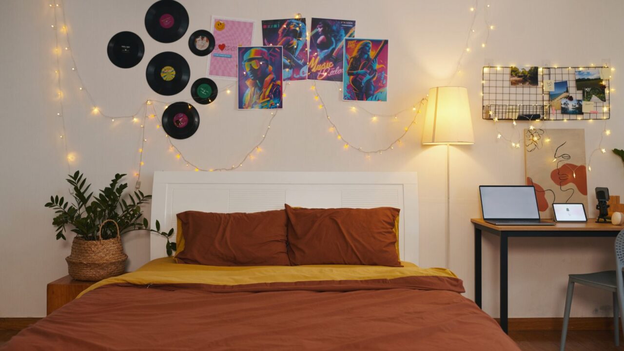 A stylish bedroom interior with a cozy bed, plants, study table, floor lamp, mesh grid, vinyl records and posters on the wall, and string lights illuminating the room.