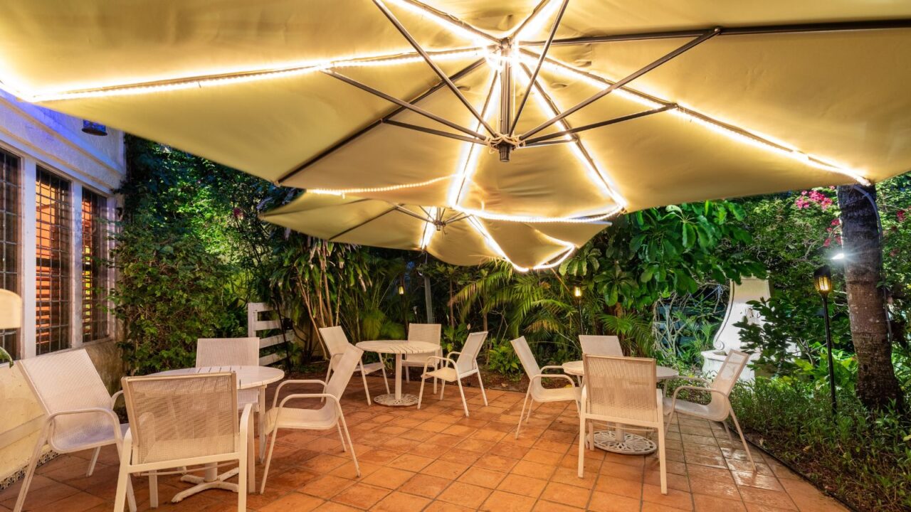 A stylish patio with tables and chairs illuminated by umbrella lights and surrounded by trees.