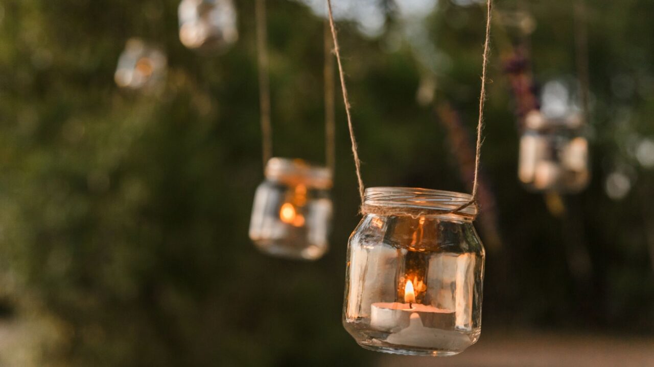 Candle jar holders hanging from trees with burning candles in them.