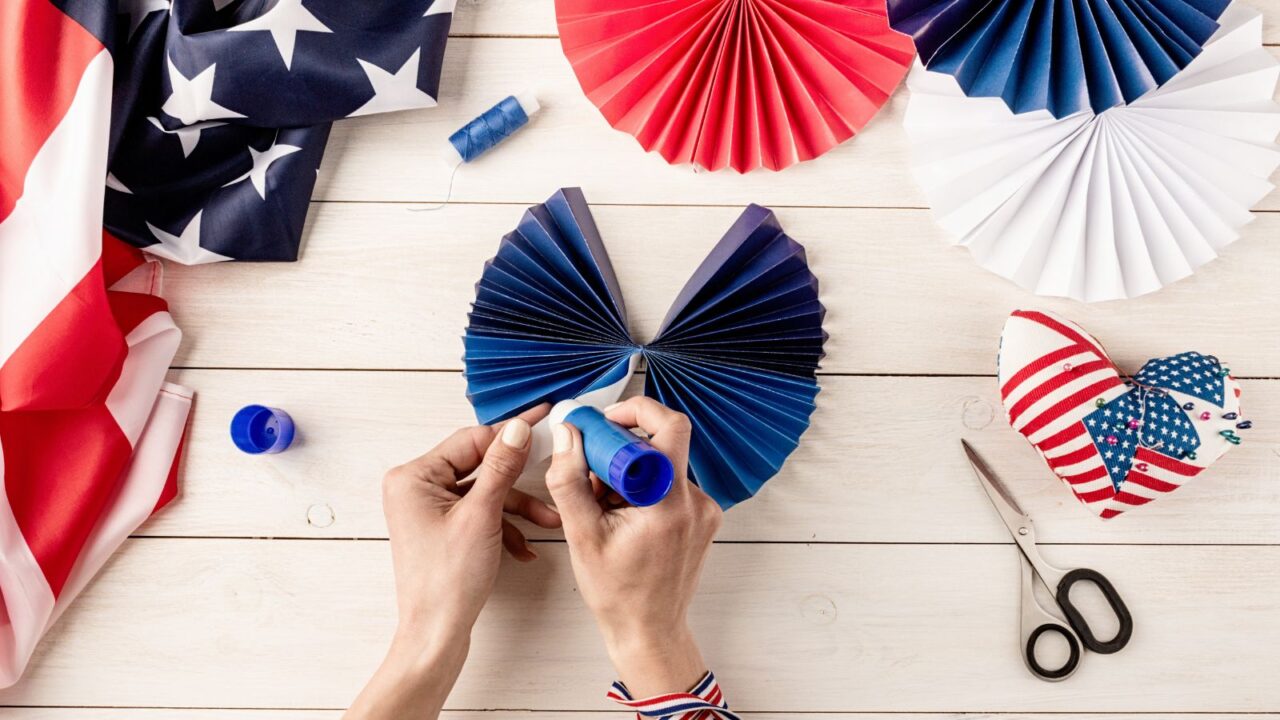 A person making USA flag themed red, blue, and white paper fans on a wooden table.