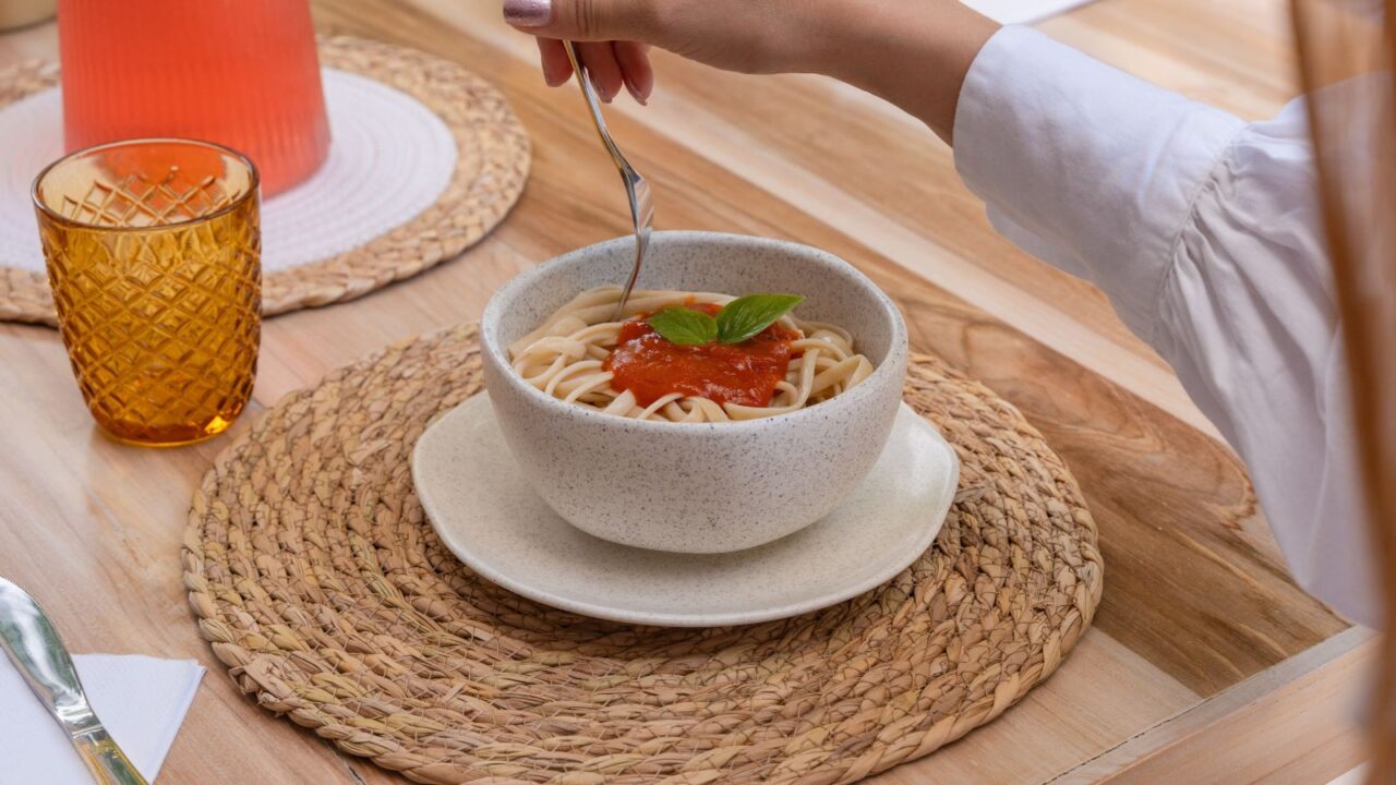 A woman holding a fork over a spaghetti bowl placed on a wicker coaster on a wooden table.