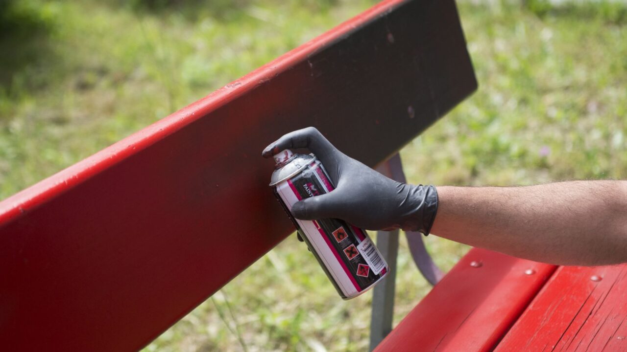 A man wearing gloves doing red spray paint on a bench.