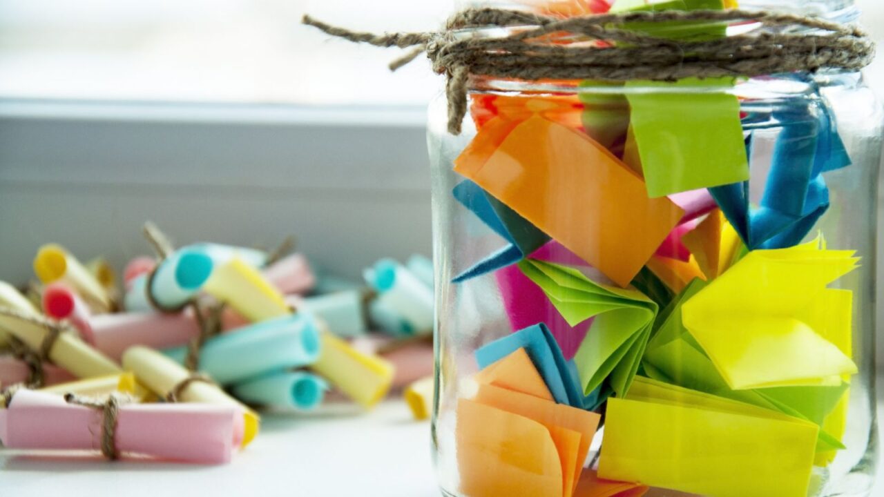 A memory jar filled with colorful notes placed on a table with small colorful messages tied with twine in the background.