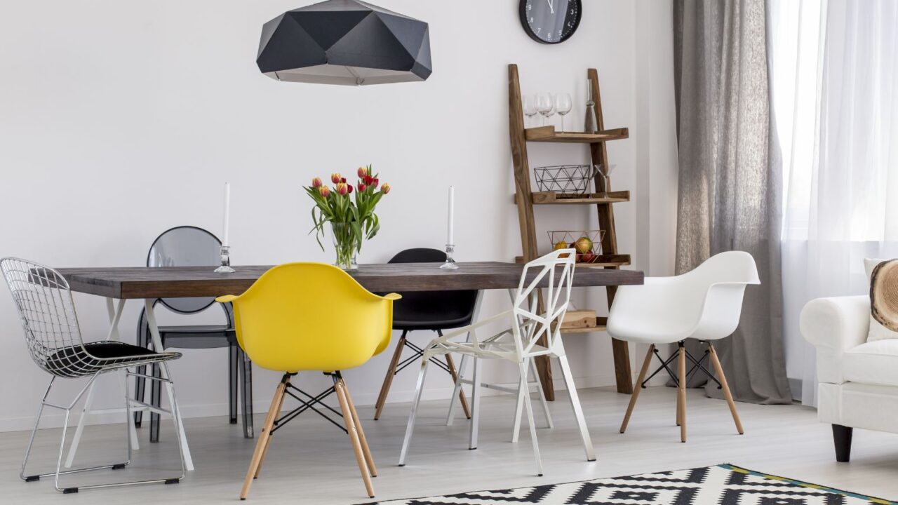 A stylish dining room interior with a wooden table, mismatched chairs, chandelier, flower vase, wooden ladder shelf, and sheer curtains.