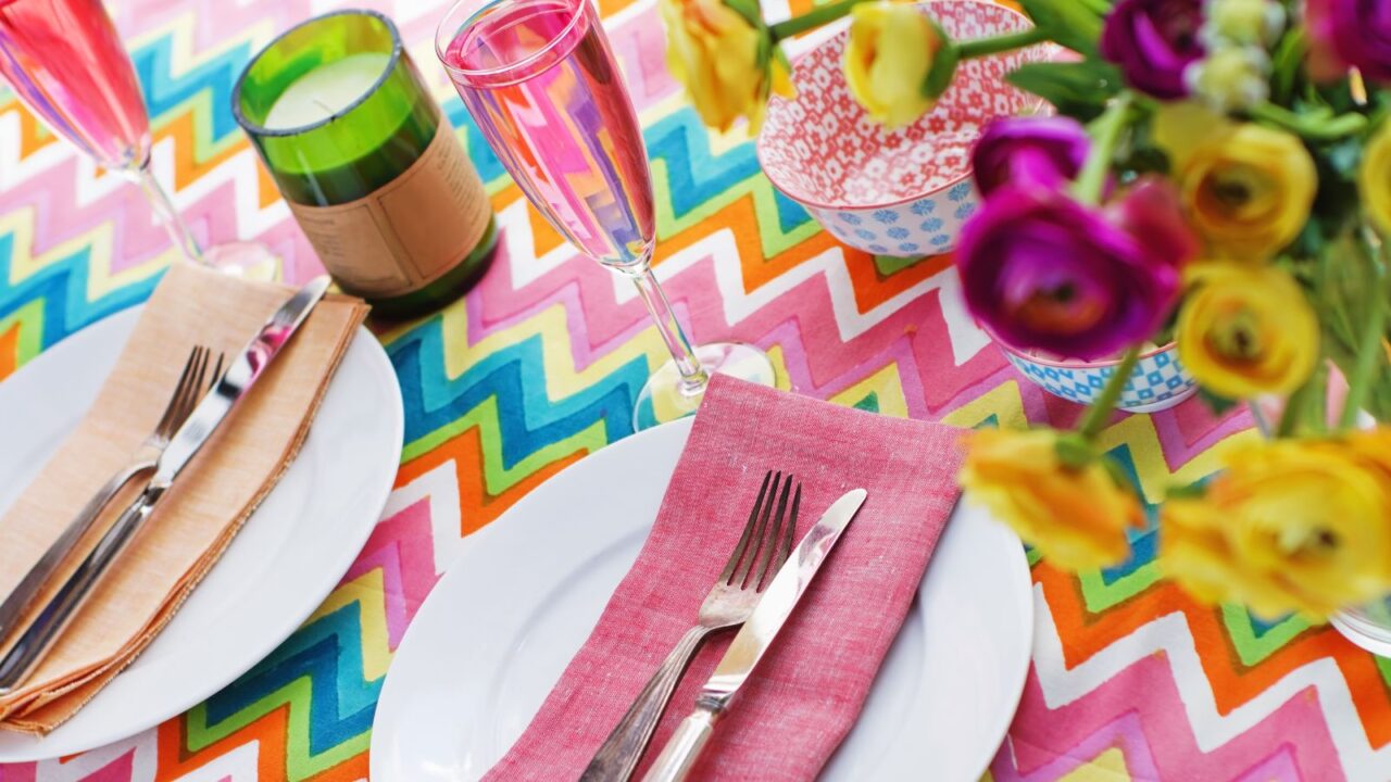 A dining table with a colorful patterned tablecloth, candle, plates, and colored napkins.
