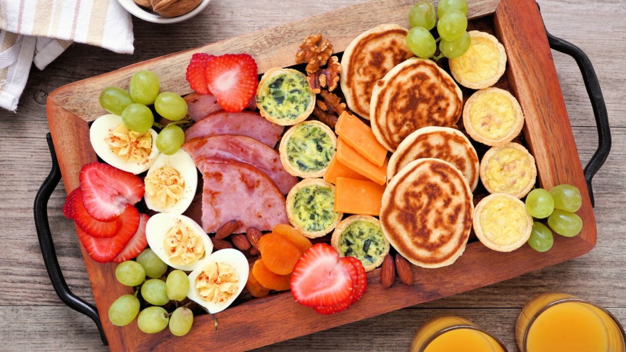A wooden grazing board with pancakes, eggs, fruits, and ham.