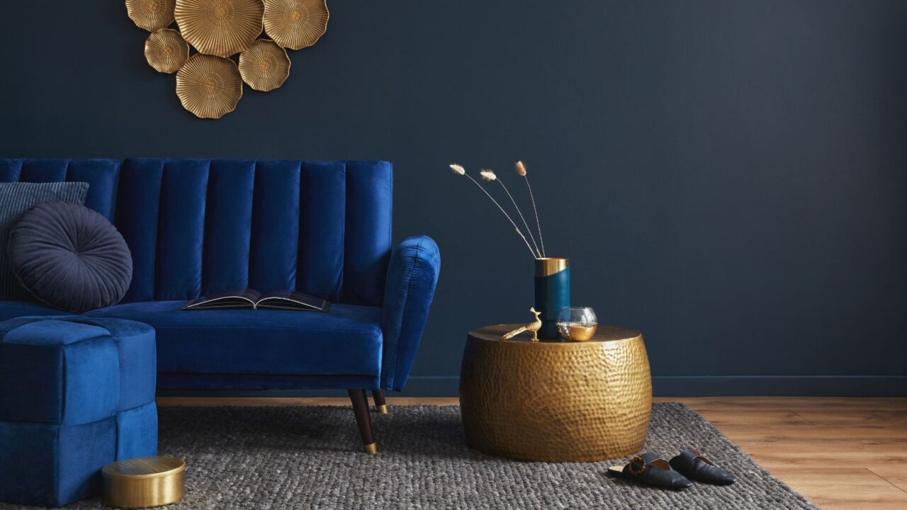 A velvet blue sofa in a living room with a dull gold metallic side table beside it with vase and sculpted decor accents.