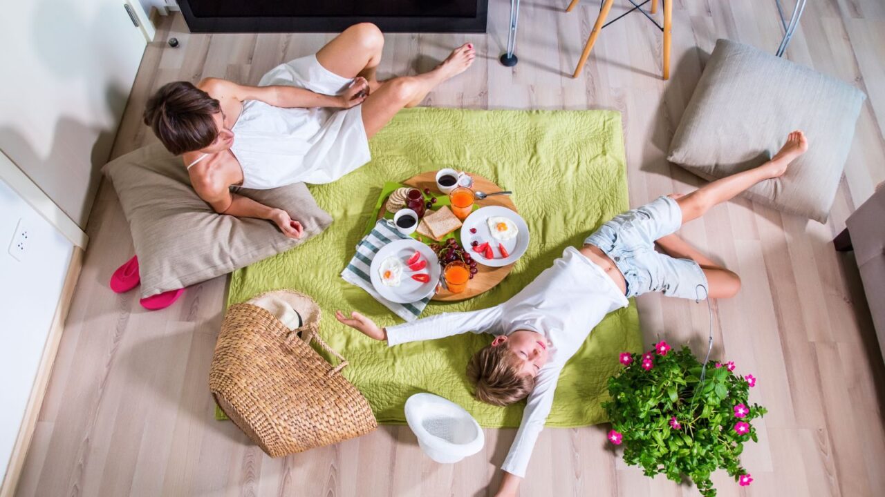 A mom and son relaxing in a cozy indoor picnic corner lying on a soft rug with flowering plant, snacks, and floor cushions near them.