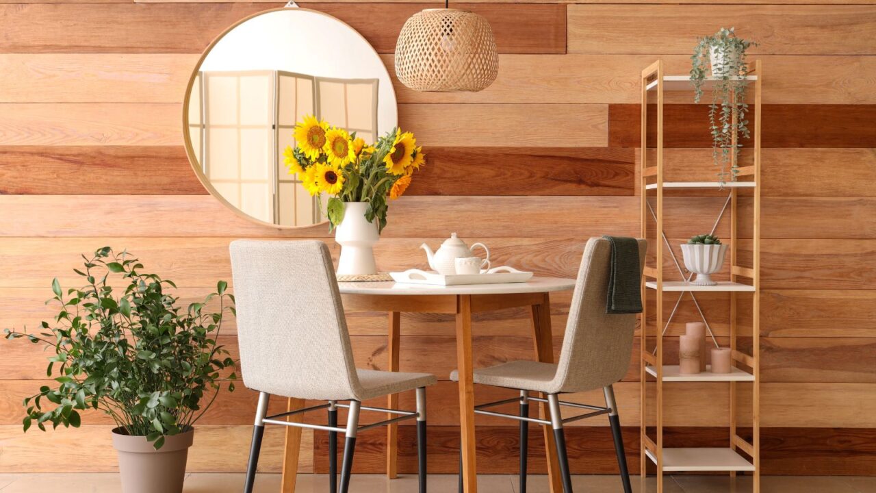 A cozy breakfast nook with a wooden table, chairs, flower vase, wooden shelf, plants, and a round mirror hanging on the wall.