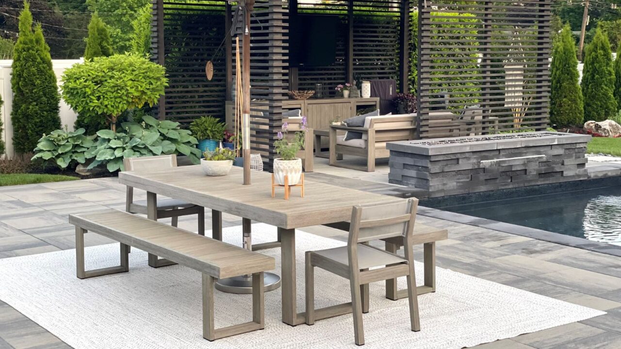An outdoor dining area with a wooden table, chairs, bench with a rug underneath, surrounded by plants.
