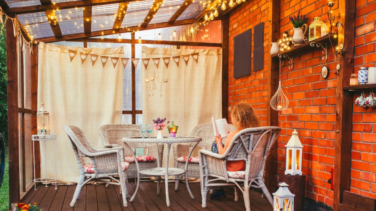 A beautiful outdoor dining setup with rattan table and chairs, curtains, string lights, and a floating shelf on a brick wall.