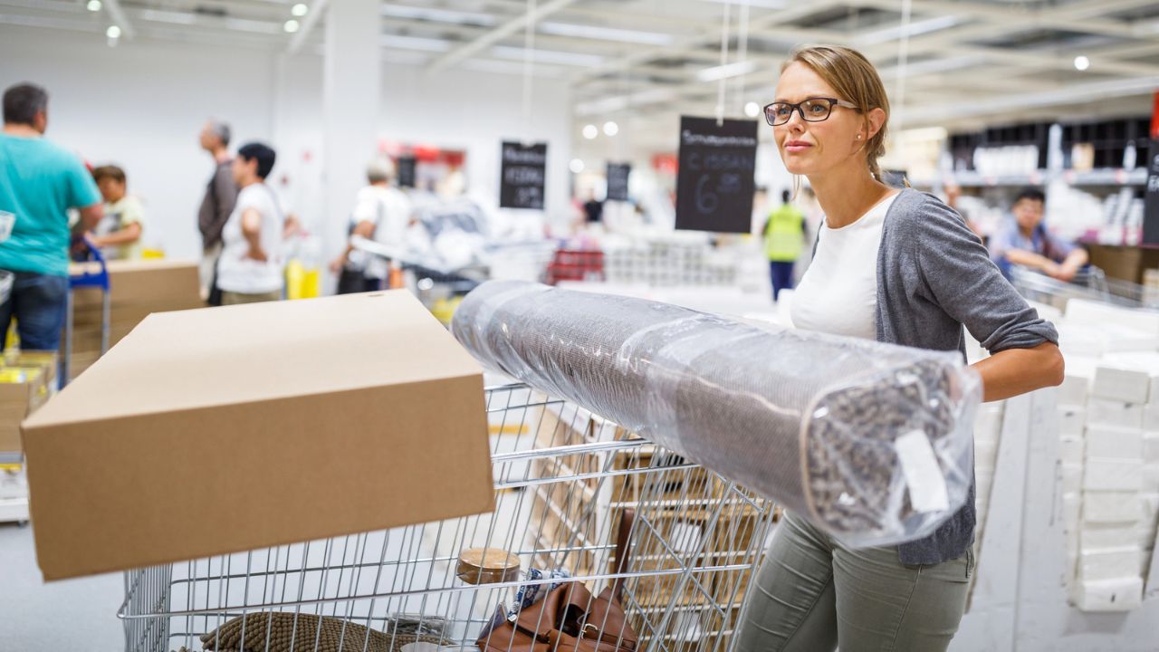 A woman shopping in a store, pushing a cart with a roll of wallpaper.