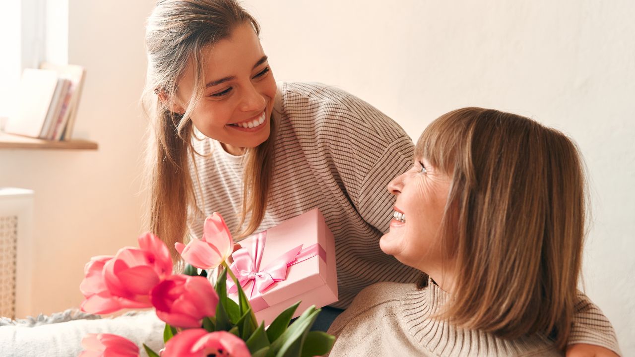 A woman giving a gift to an older woman with flowers; they are smiling.