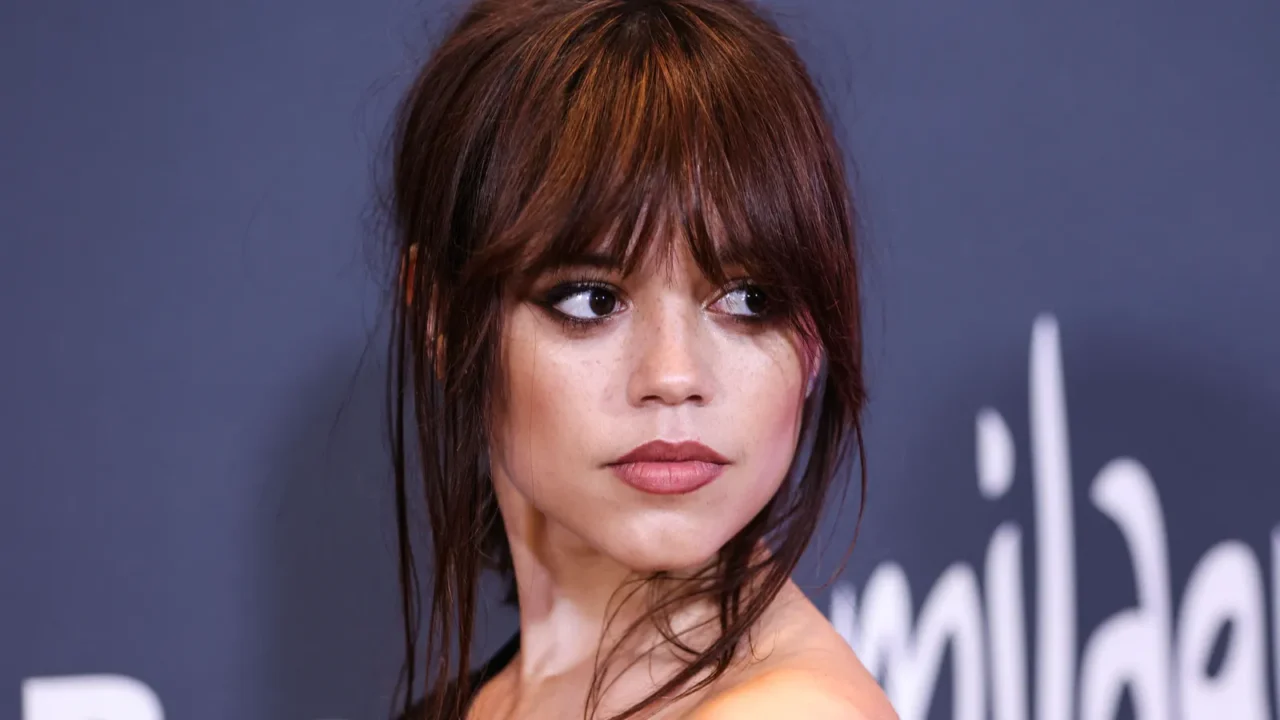 A close-up of Jenna Ortega with tousled hair, subtle makeup, and a contemplative expression against a dark background.