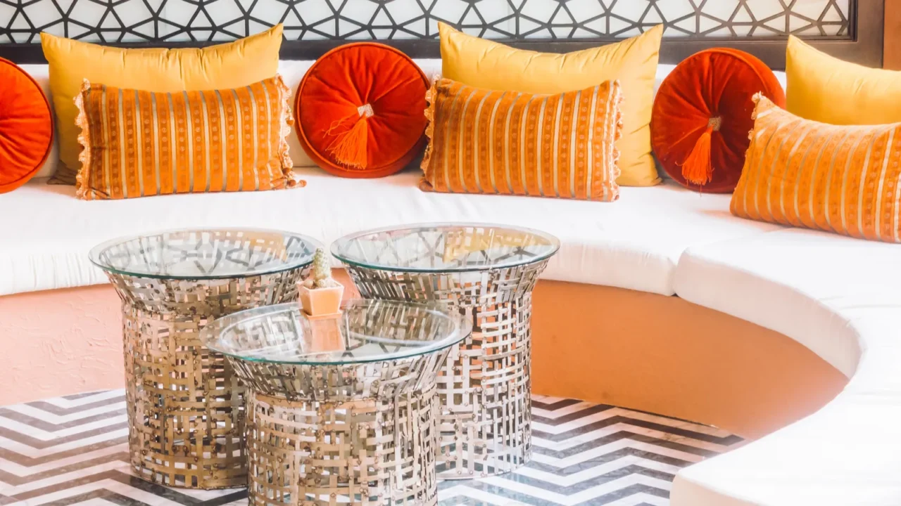 A stylish living room corner with a white curved sofa, orange and yellow cushions, glass-top metallic tables, and a black-and-white zigzag patterned rug.