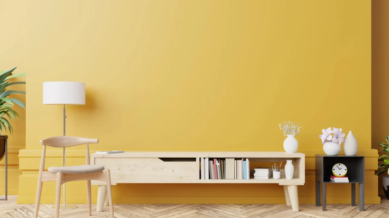 A living room interior with a wooden armchair, floor lamp, plants, and a wooden console on a wooden floor in front of a golden wall.