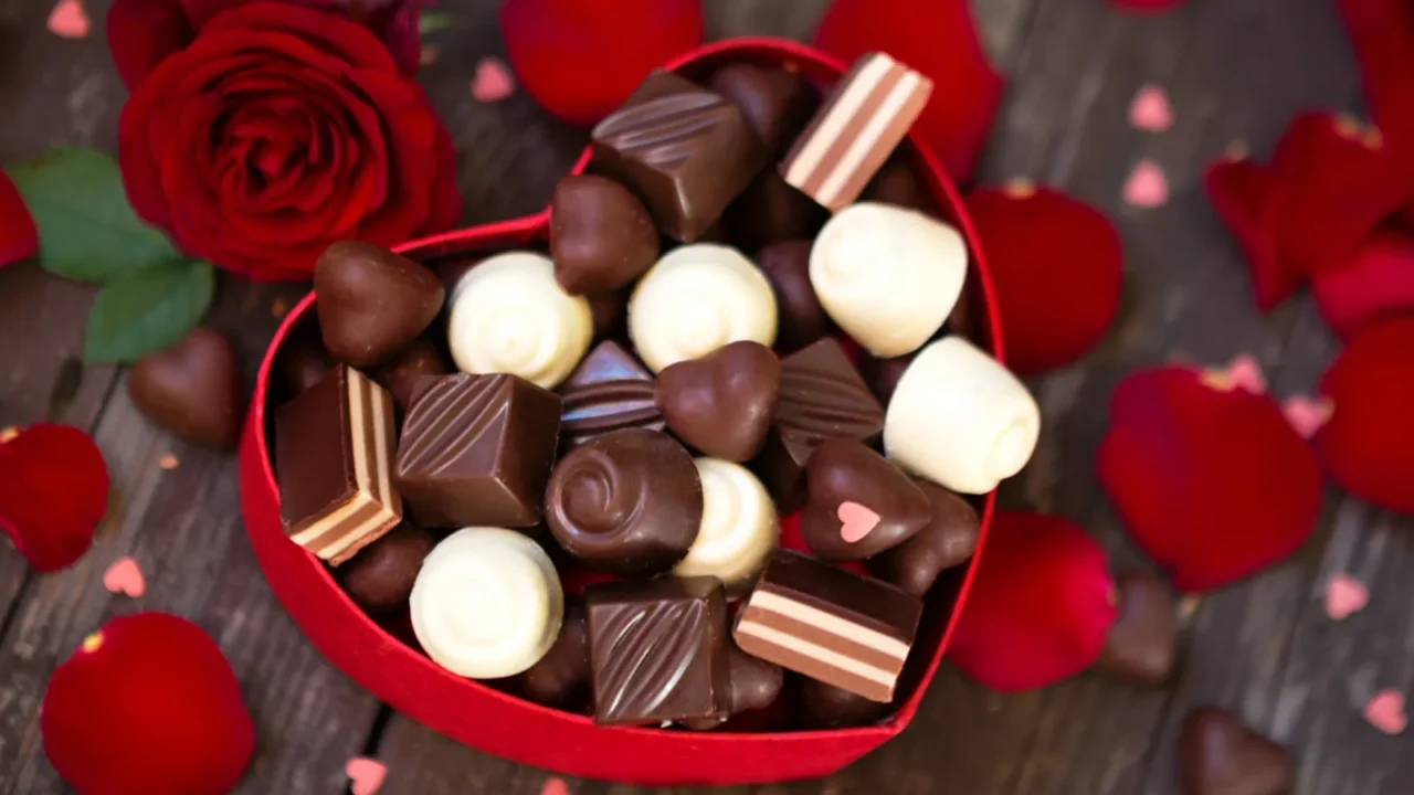 A heart-shaped box of assorted chocolates surrounded by red rose petals.