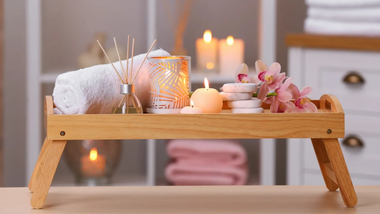 A wooden bathroom tray with a folded towel, flowers, candles, and stones.