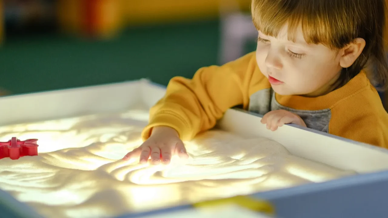A small boy playing at a sensory bin station with a light table filled with sand.