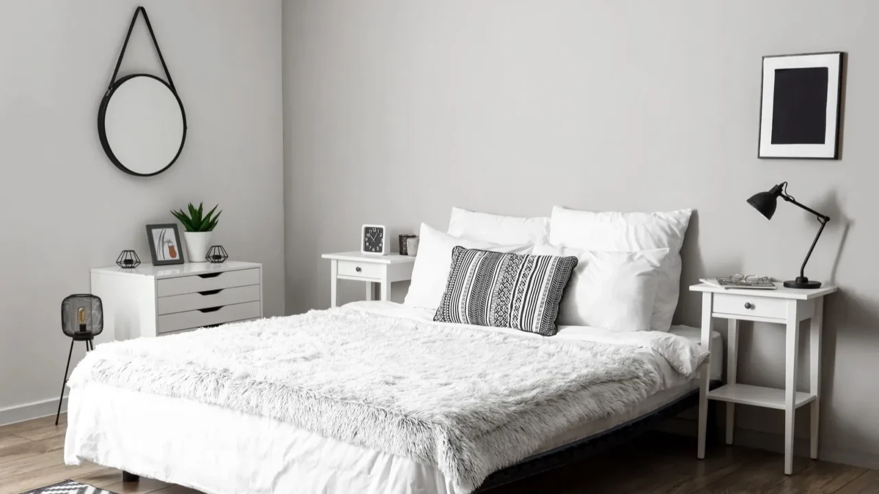 A white bedroom interior with cozy bed, nightstands, round hanging mirror, and white shelf with drawers and decor accents on it.
