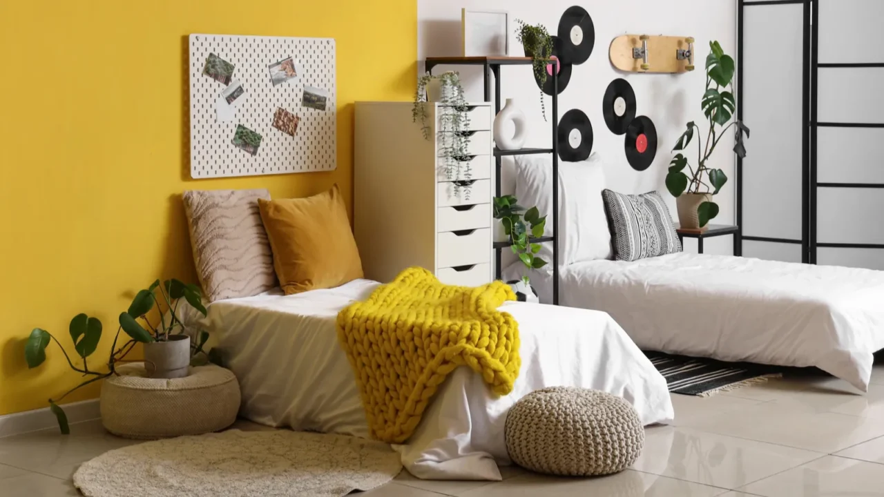 A shared bedroom interior with two single beds, storage shelf, floor cushion, and a pegboard on the yellow wall with pictures on it.