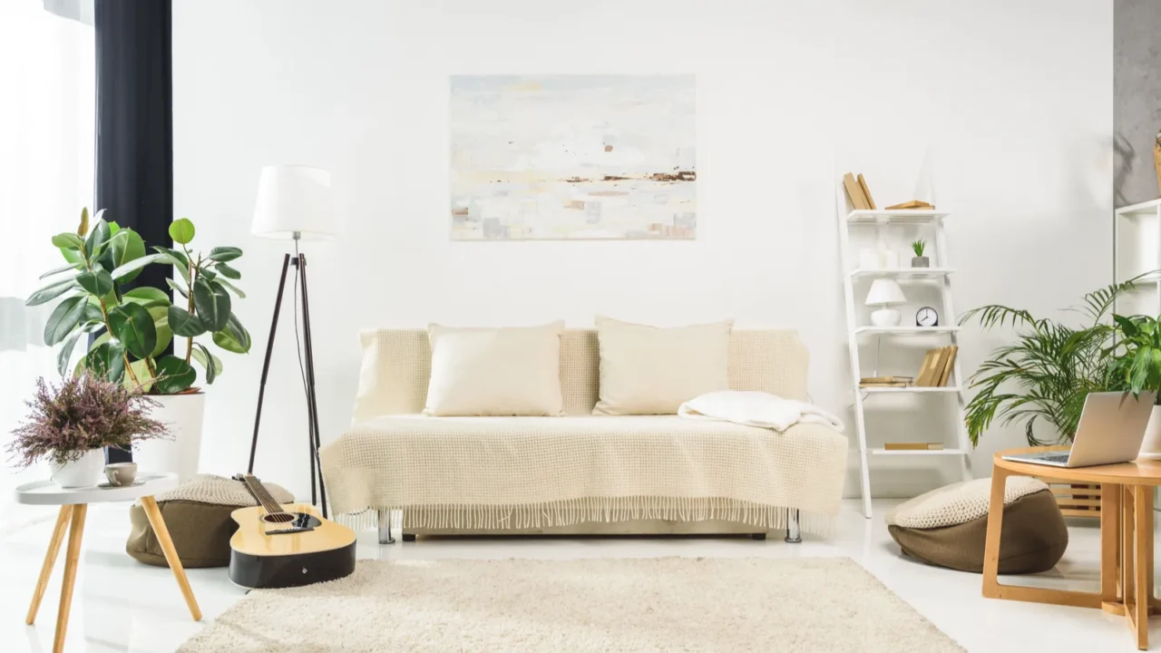 A stylish living room interior with a cozy sofa, floor lamp, ladder shelf, guitar, rug, plants, and natural light coming through the window.