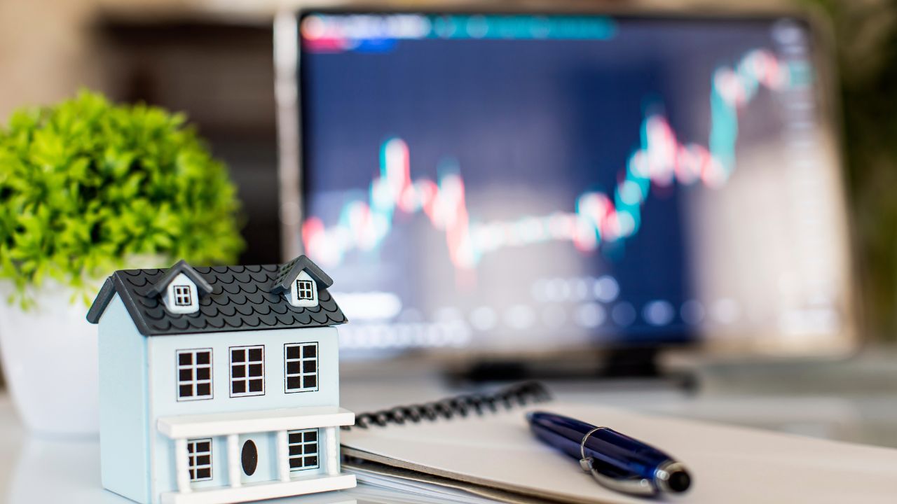 A small decorative house with a laptop showing stock market graphs nearby.