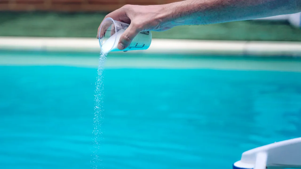 Hand adding a chemical or chlorine tablet to the pool water.