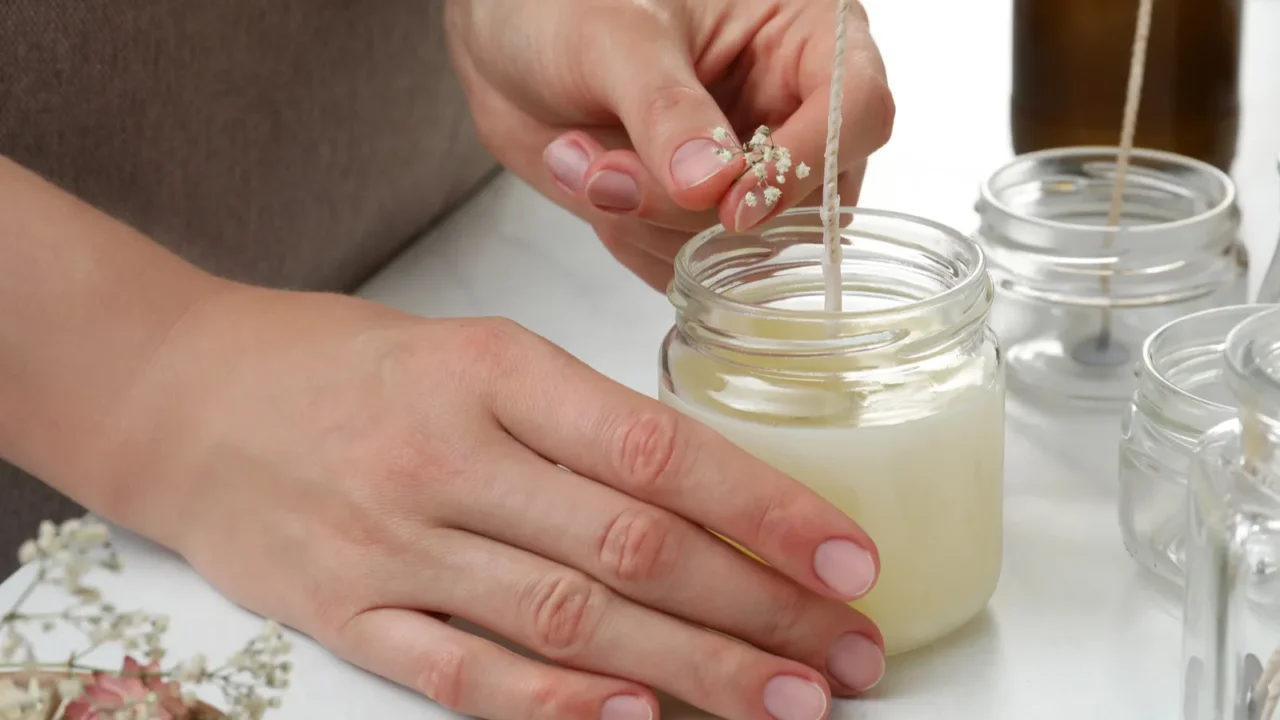 A woman making candle in a glass jar kept on a white table.
