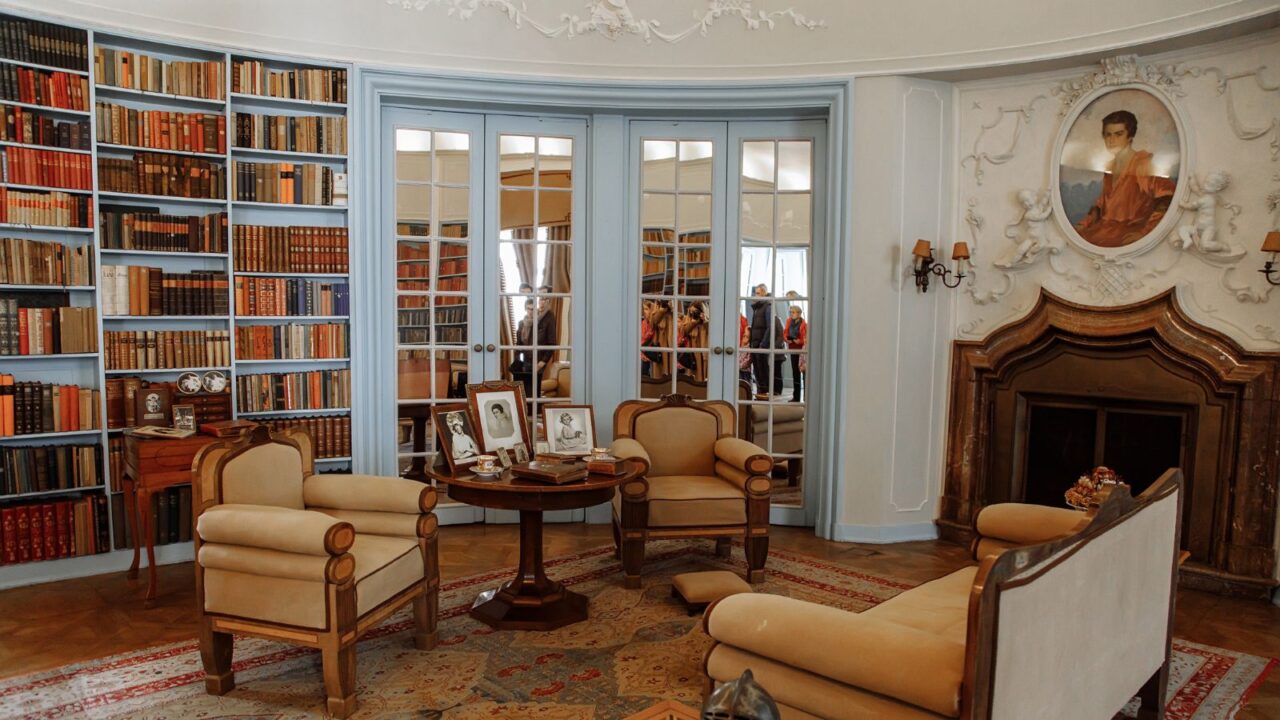 A cozy room with a light blue bookshelf wall filled with books, antique-style chairs, and framed photographs, with a fireplace and a large mirror reflecting the interior.
