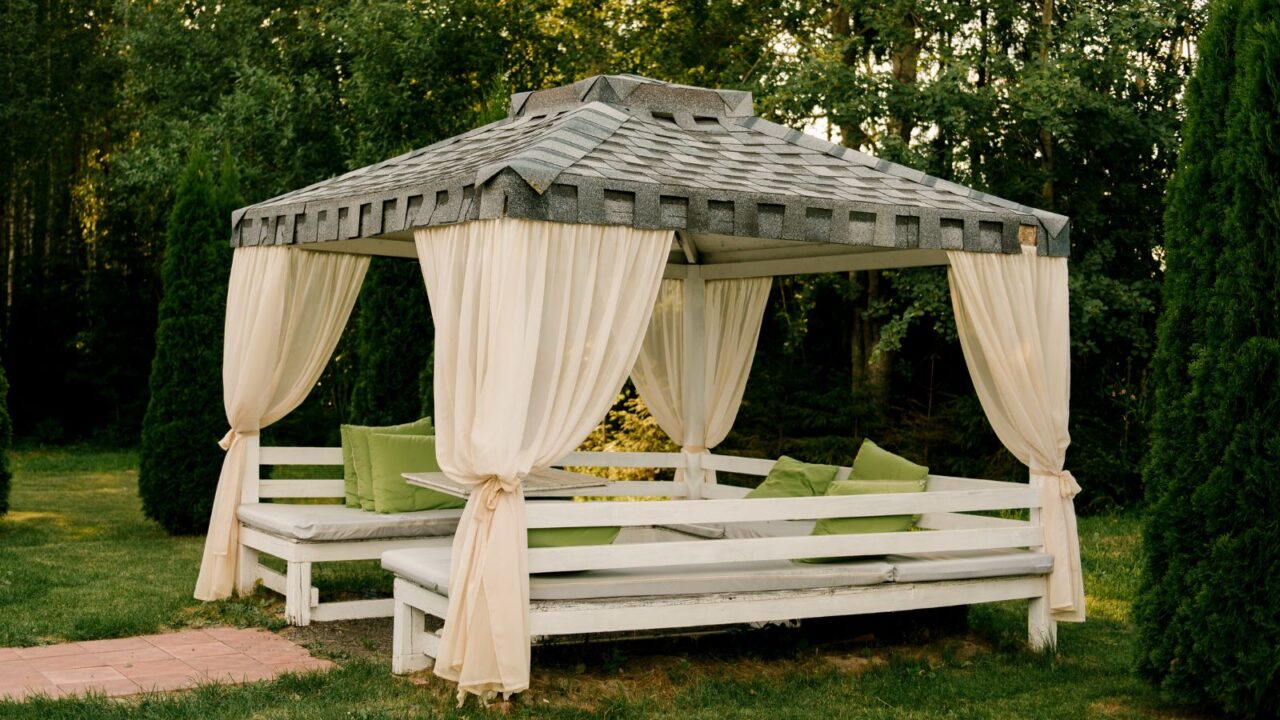 An outdoor gazebo with draped curtains and cushions, set in a garden.