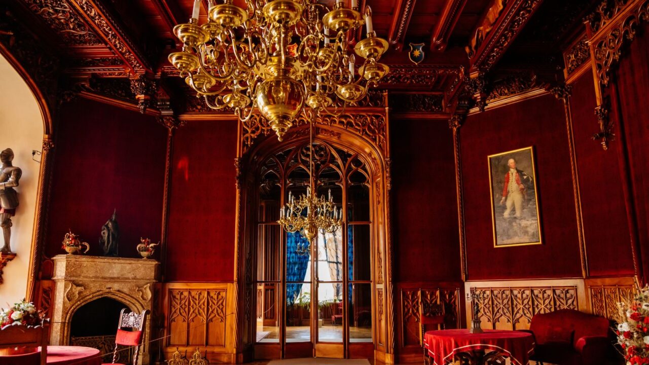 An opulent room with deep red walls, intricate woodwork, a large gold chandelier, and elegant furniture, including a fireplace and a portrait.