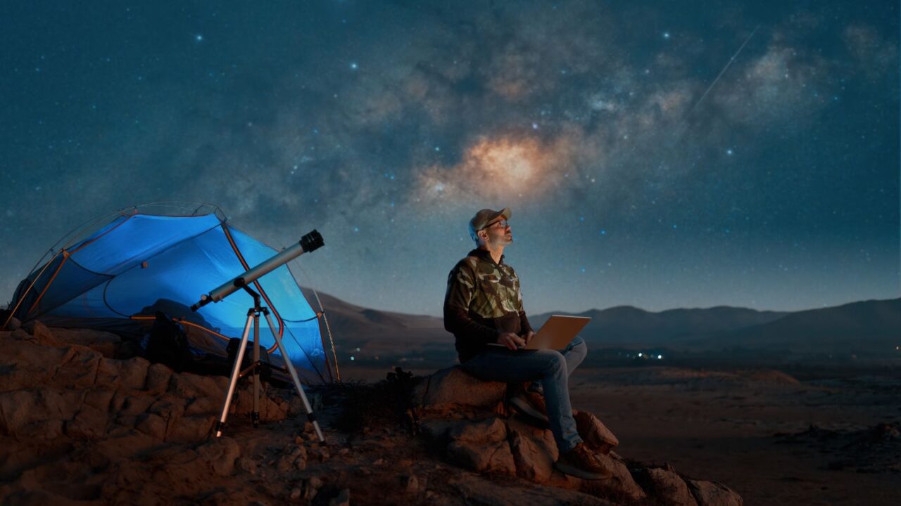 A digital nomad man sitting alone with a laptop in the desert at night next to a telescope watching the starry night.