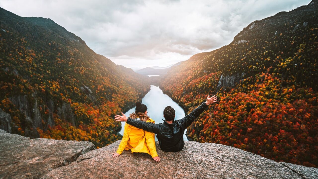 couple enjoying autumn fall foliage at indian head, adirondacks, sitting together with arms raised on a mountaintop at sunset during a hiking adventure