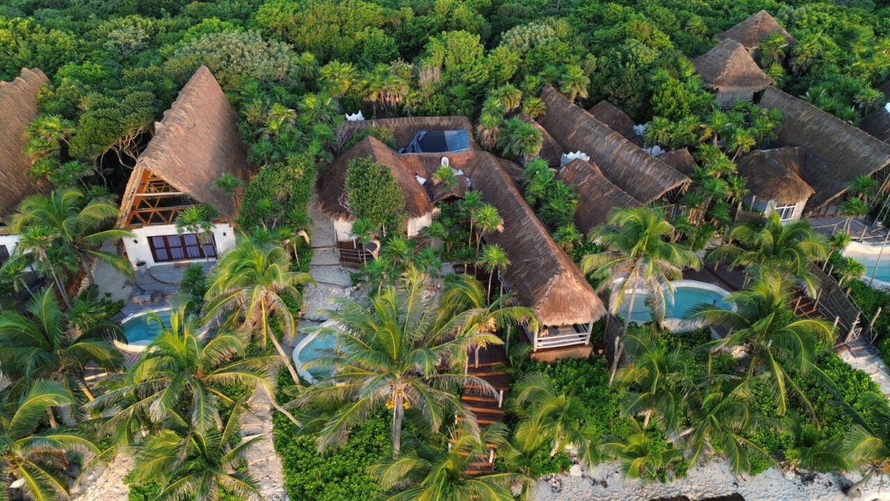 A lush tropical resort with thatched-roof bungalows and swimming pools surrounded by dense green foliage at Tulum Hotels.