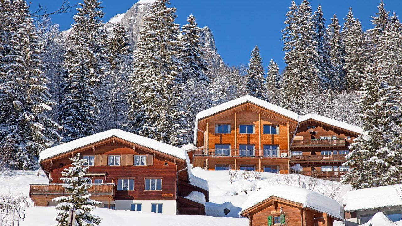 A snowy mountain landscape with wooden chalets nestled among tall snow-covered pine trees under a clear blue sky.