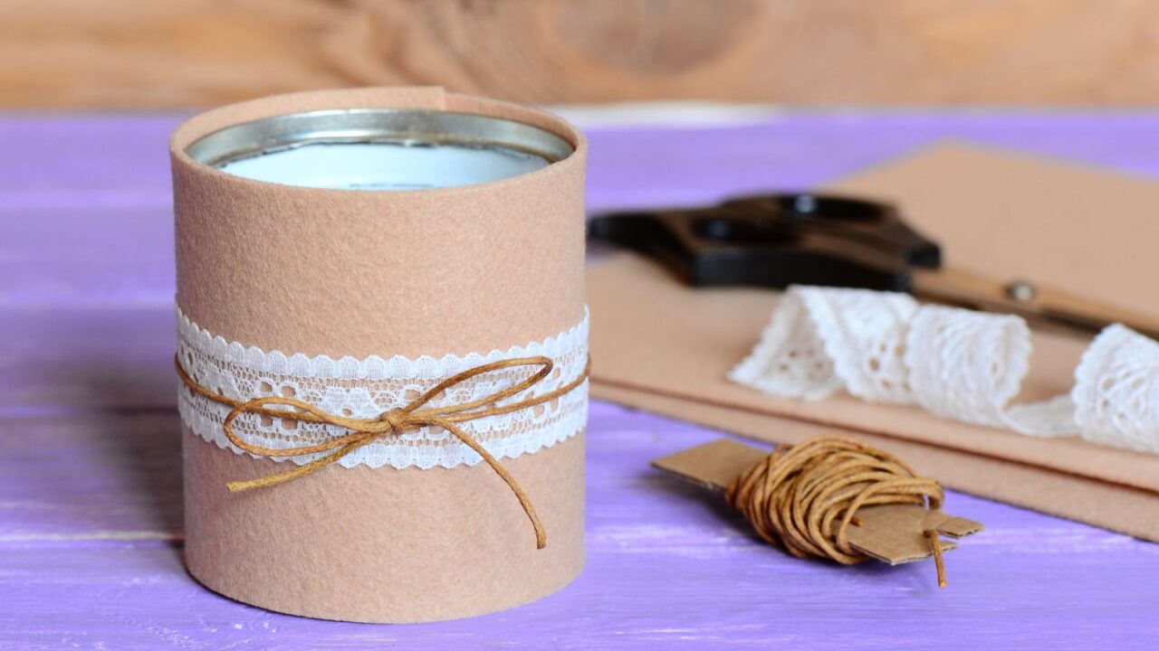 A cylindrical craft container wrapped with pink paper, lace, and twine, placed on a purple surface with crafting materials nearby.