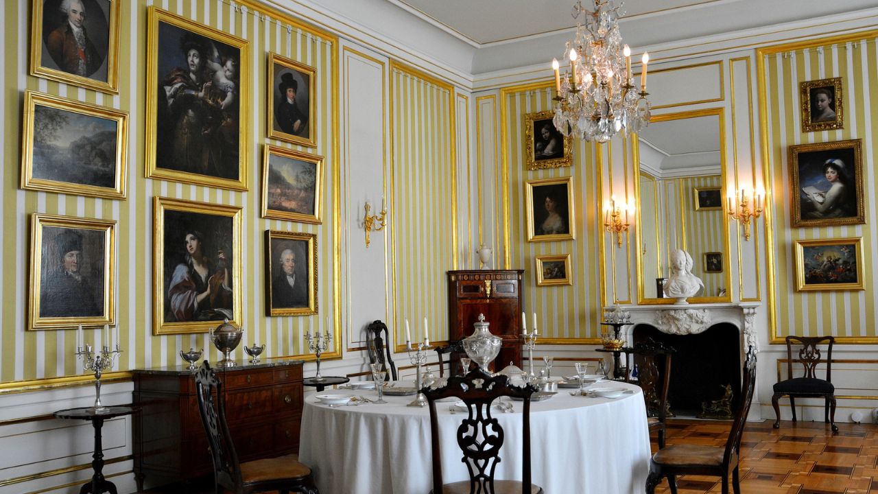 An opulent dining room with a round table, classic artwork, chandelier, and yellow-striped wallpaper.