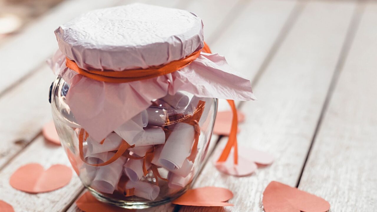 A glass jar filled with crumpled paper and pink paper hearts scattered around on a wooden surface.