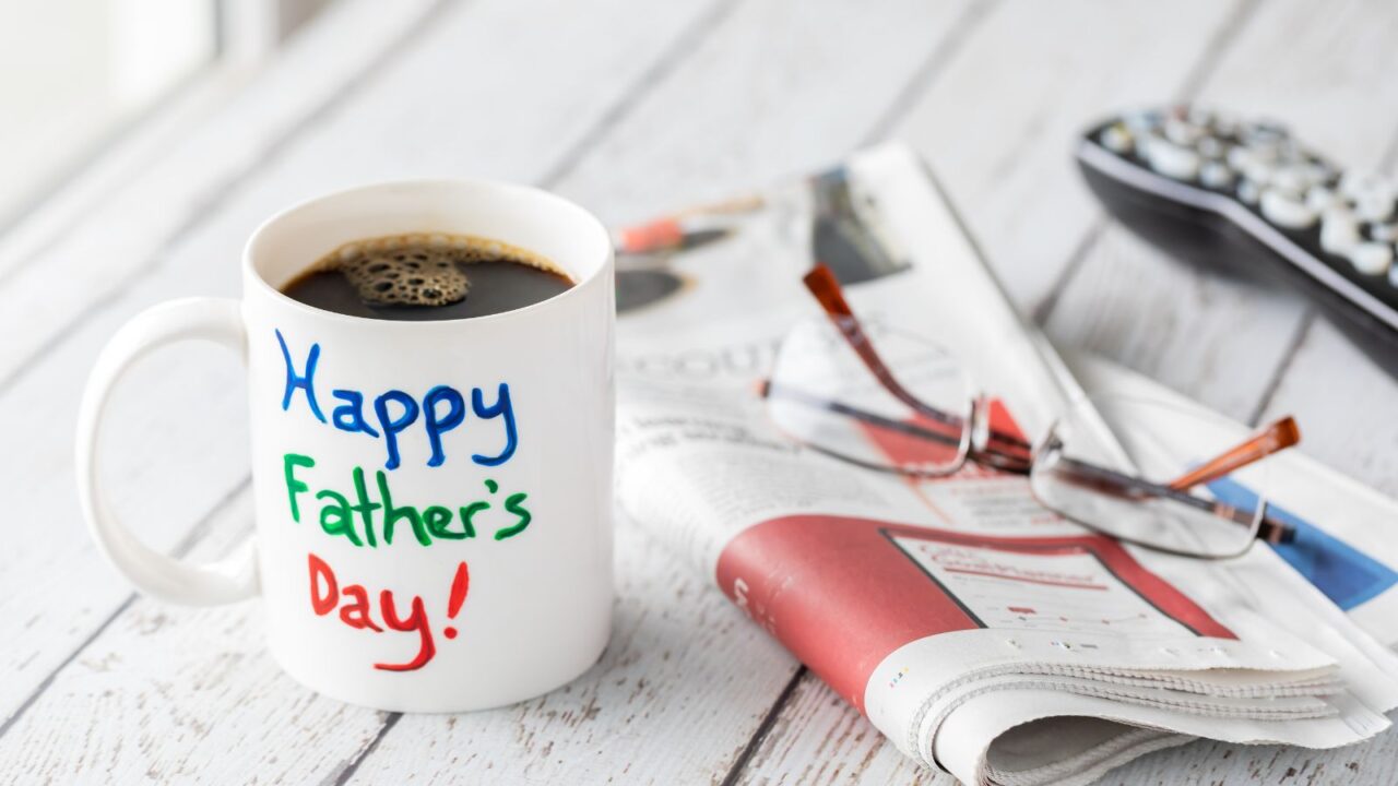 A coffee mug with "Happy Father's Day!" written on it, placed on a white wooden surface with a newspaper and glasses.